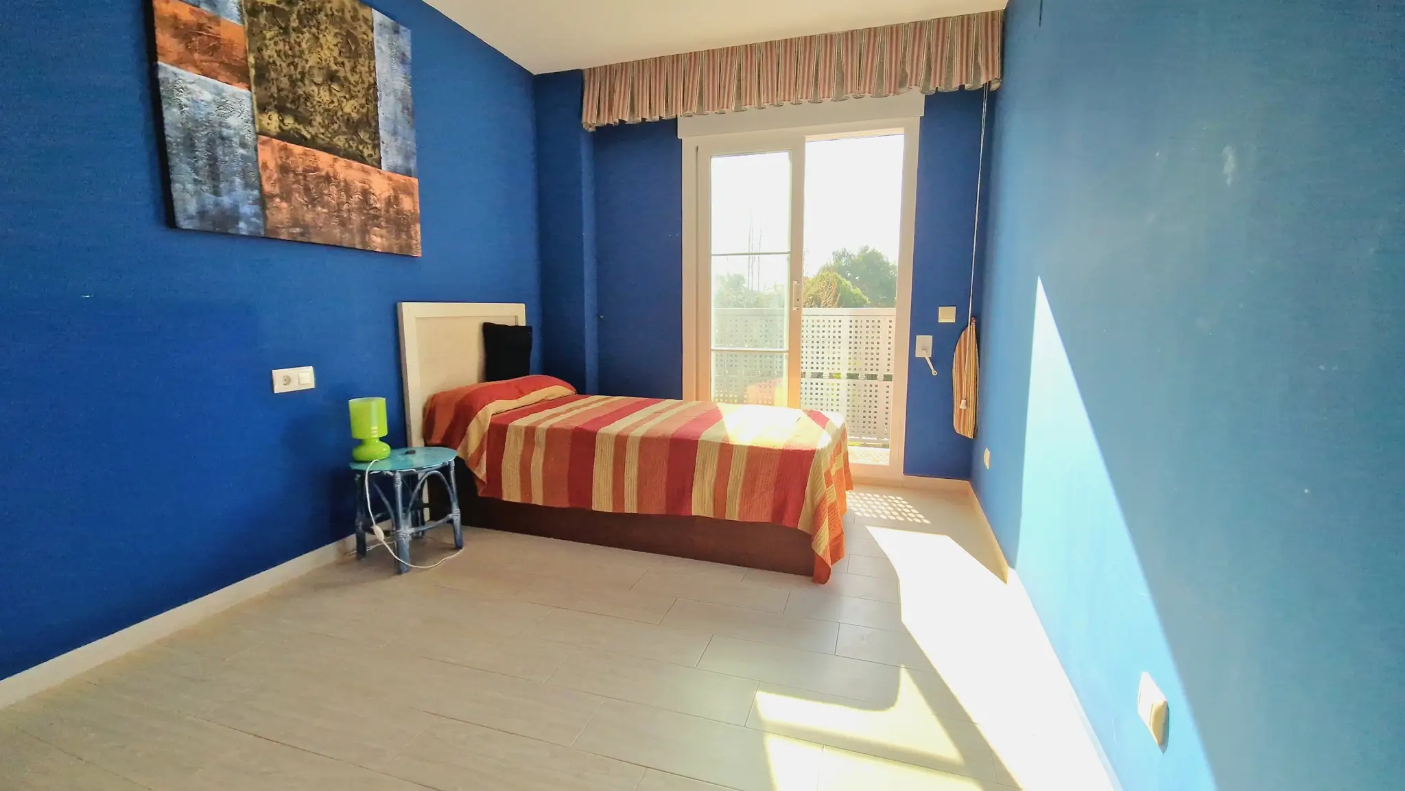 Chambre aux murs bleu profond, lit simple avec literie rayée et accès direct à un balcon par des portes vitrées coulissantes.