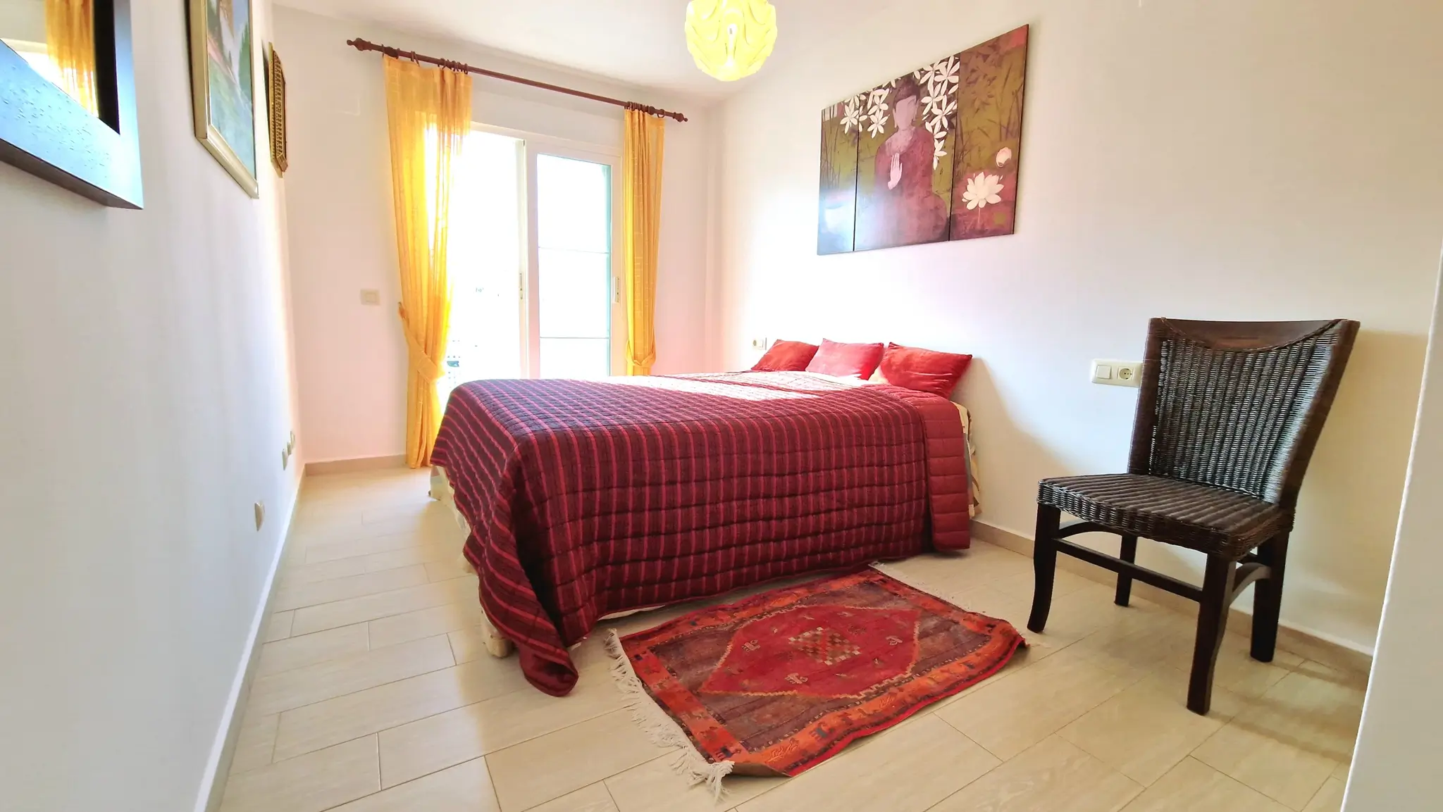Chambre lumineuse avec un lit double, couvre-lit rouge matelassé, rideaux jaunes, chaise en osier foncé et un triptique décoratif de Bouddha au mur.