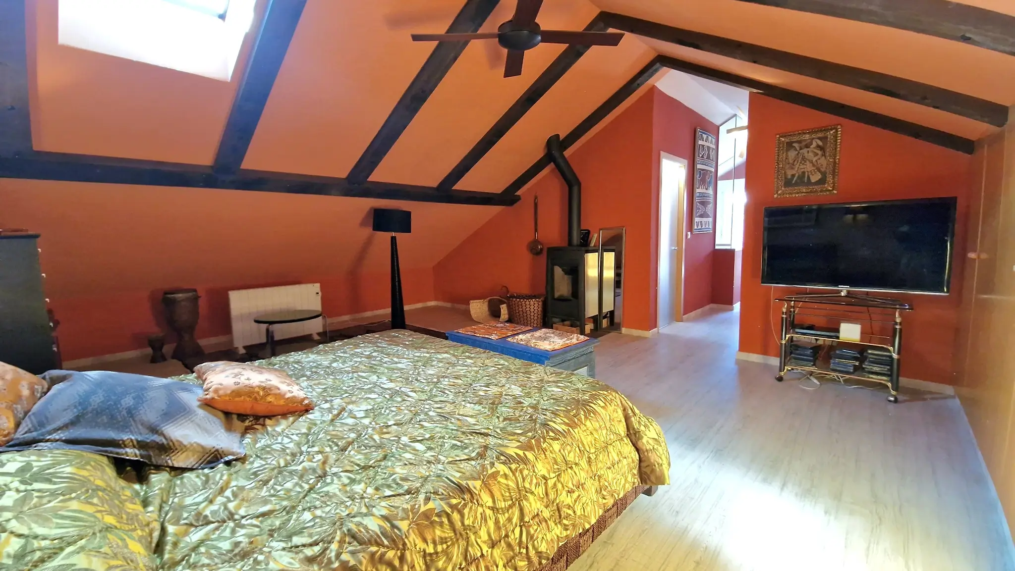 Chambre mansardée avec poutres apparentes, murs terracotta, poêle à bois et lucarne offrant une luminosité naturelle sur le parquet.