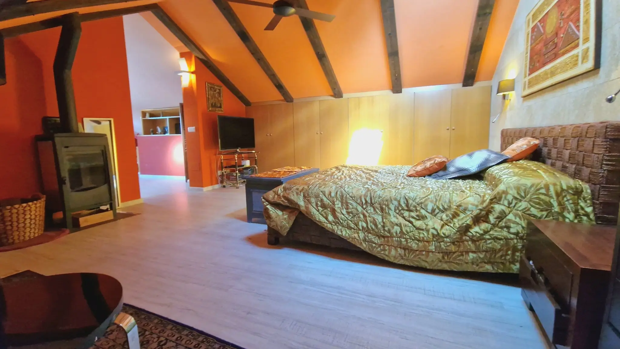 Chambre mansardée avec poutres en bois apparentes, poêle à bois et murs orange. Dispose de placards intégrés, d'un ventilateur de plafond et d'une tête de lit tressée.