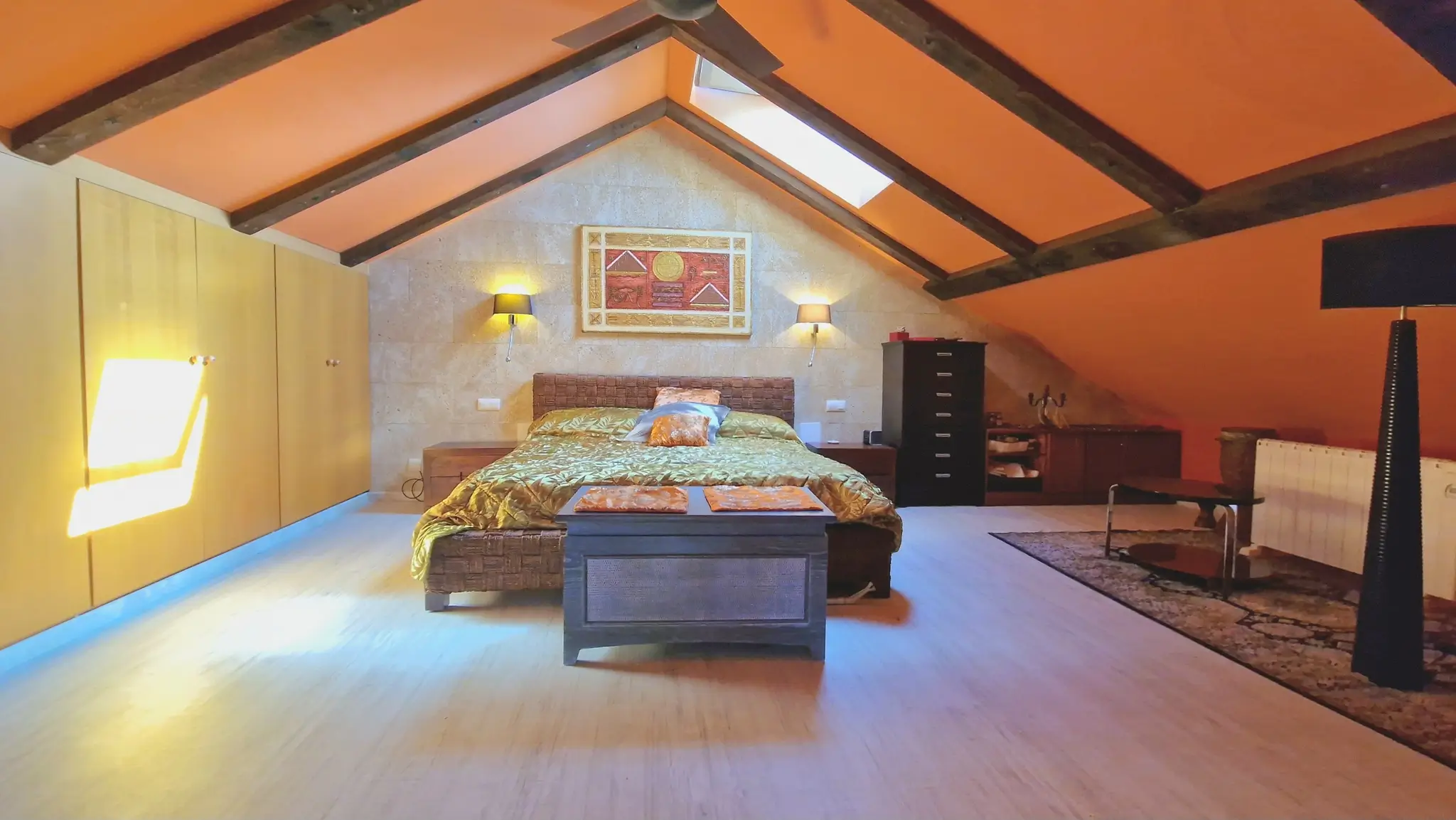 Chambre mansardée avec poutres en bois sombre, plafonds inclinés orange, mur d'accent en pierre et grands placards intégrés en bois clair.
