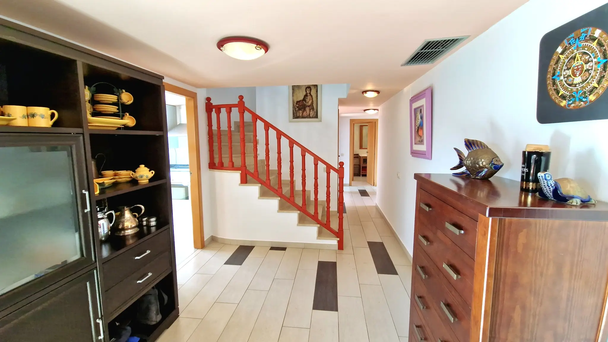 Couloir avec escalier à rampe en bois rouge, sol carrelé avec accents sombres, vitrine en bois foncé et commode agrémentée d'objets décoratifs.