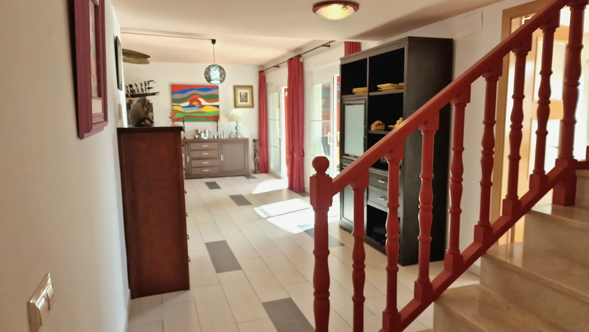 Couloir avec rampe en bois rouge et escalier en marbre, menant à un espace de vie lumineux avec carrelage au sol, meubles sombres et rideaux rouges.