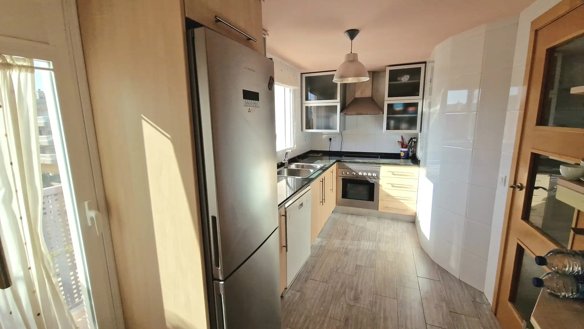 Cuisine avec meubles en bois clair, plans de travail en granit noir et appareils en acier inoxydable. Sol en carrelage imitation bois et luminosité naturelle.