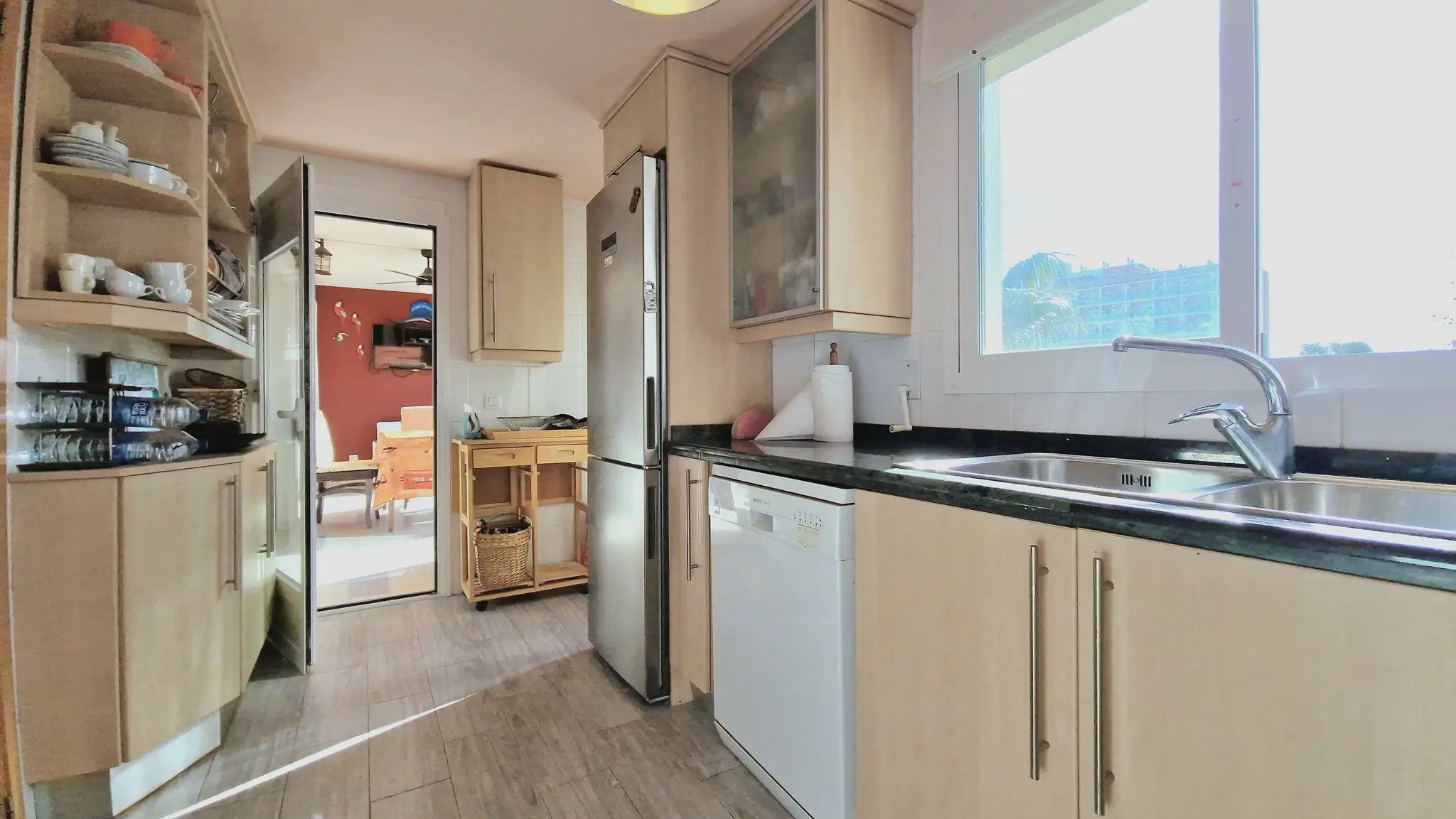 Cuisine fonctionnelle avec meubles en bois clair, plan de travail en granit sombre, évier en inox et vue dégagée par la fenêtre.