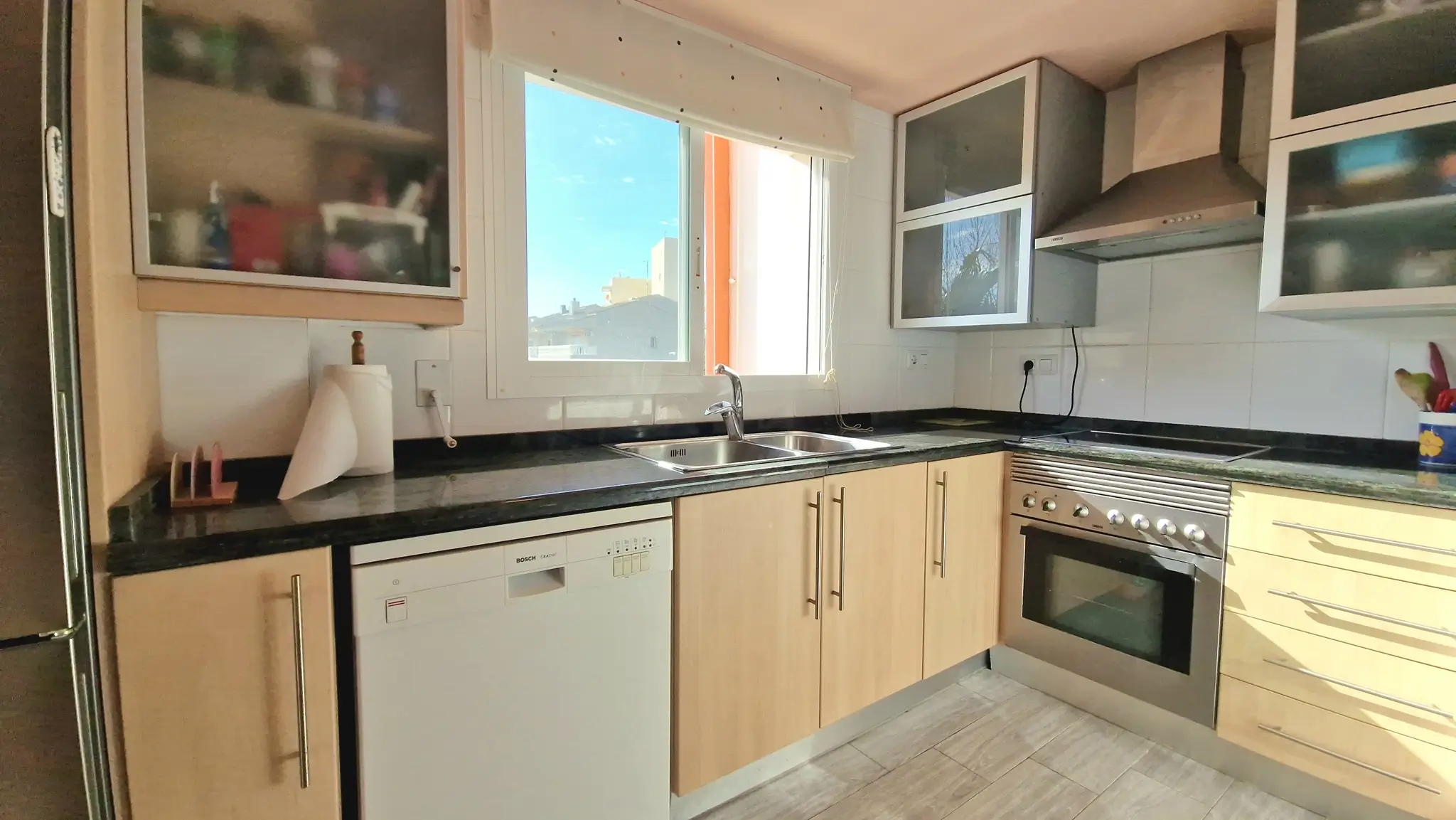 Cuisine fonctionnelle avec placards en bois clair, plan de travail en granit sombre et électroménager intégré dont un lave-vaisselle Bosch et un four en inox.
