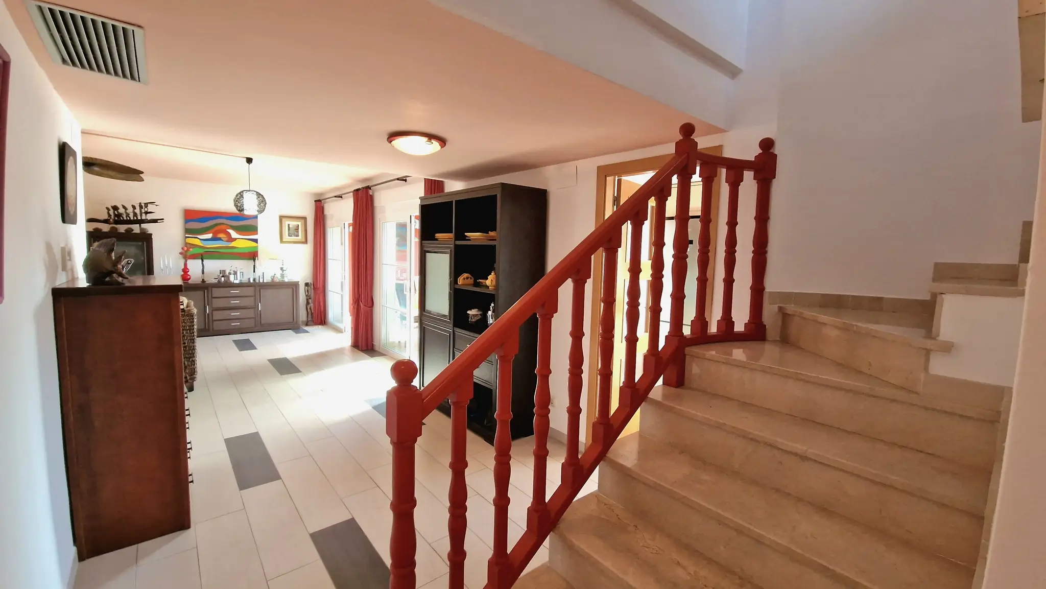 Escalier en marbre avec rampe en bois rouge ouvrant sur un salon lumineux doté de carrelage bicolore et de meubles en bois sombre.