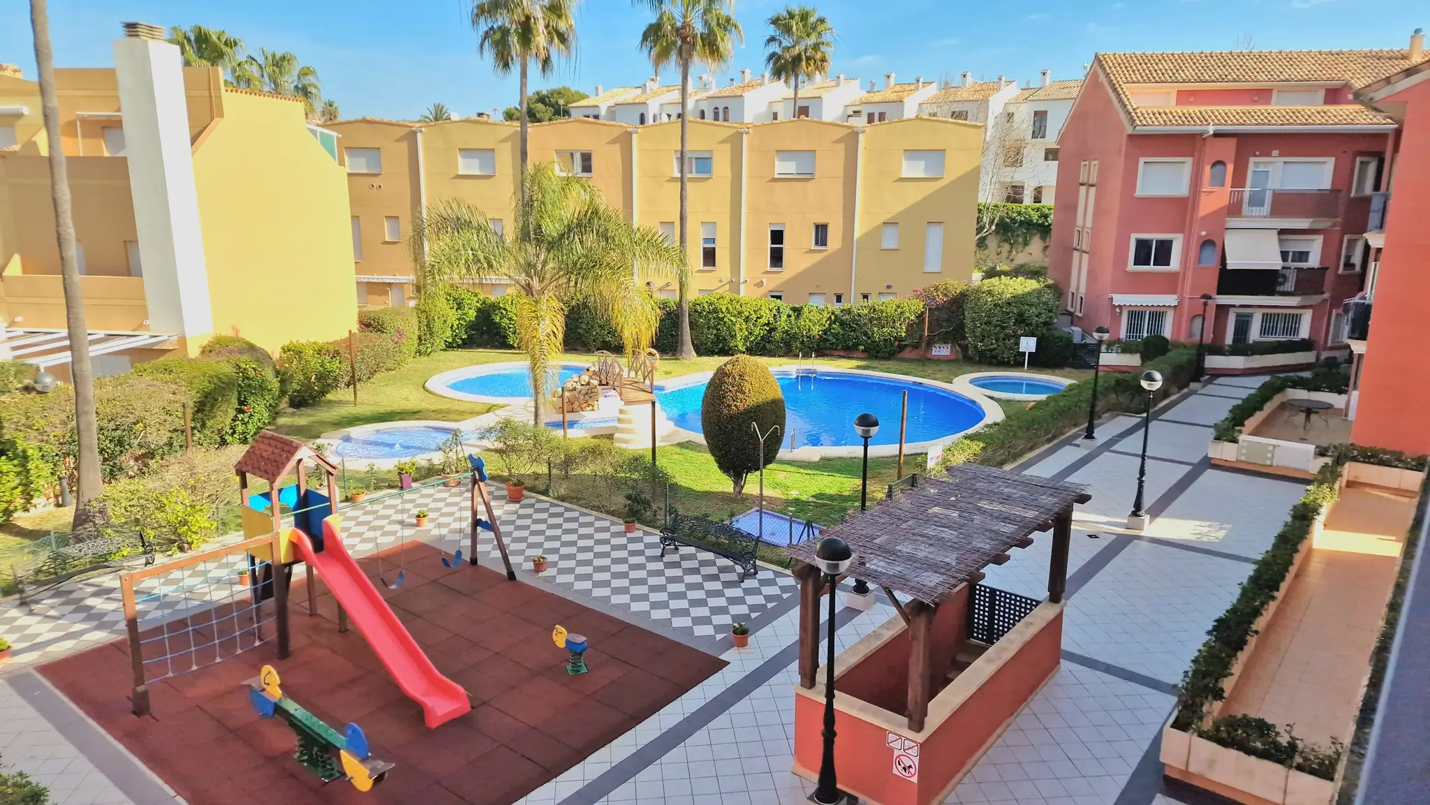 Espace commun comprenant une aire de jeux, une piscine avec pont et des jardins paysagers, entouré de résidences aux façades ocres et jaunes sous un ciel clair.