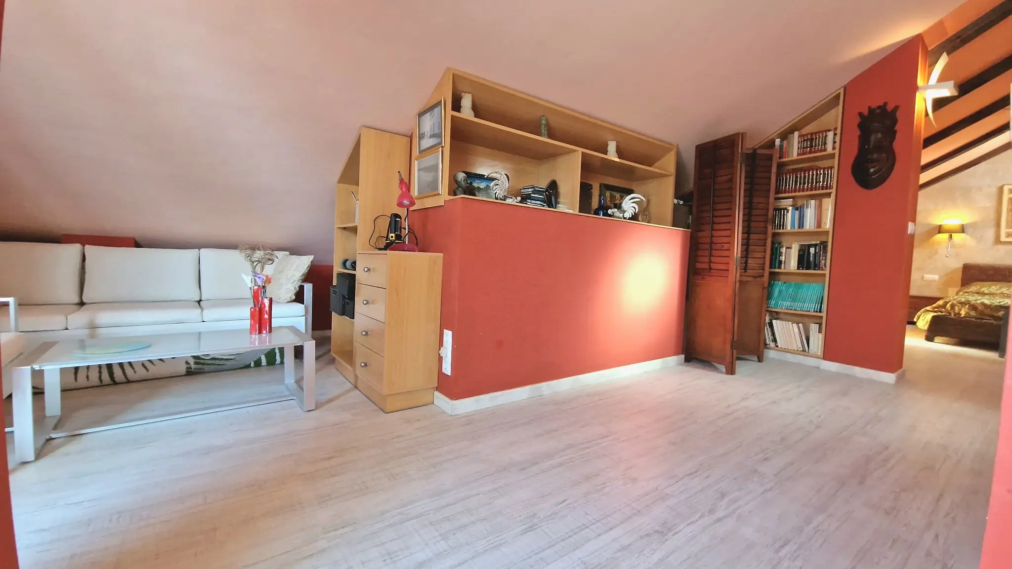 Espace de vie mansardé avec des murs terracotta, des étagères en bois intégrées et un sol stratifié clair sous plafond incliné.