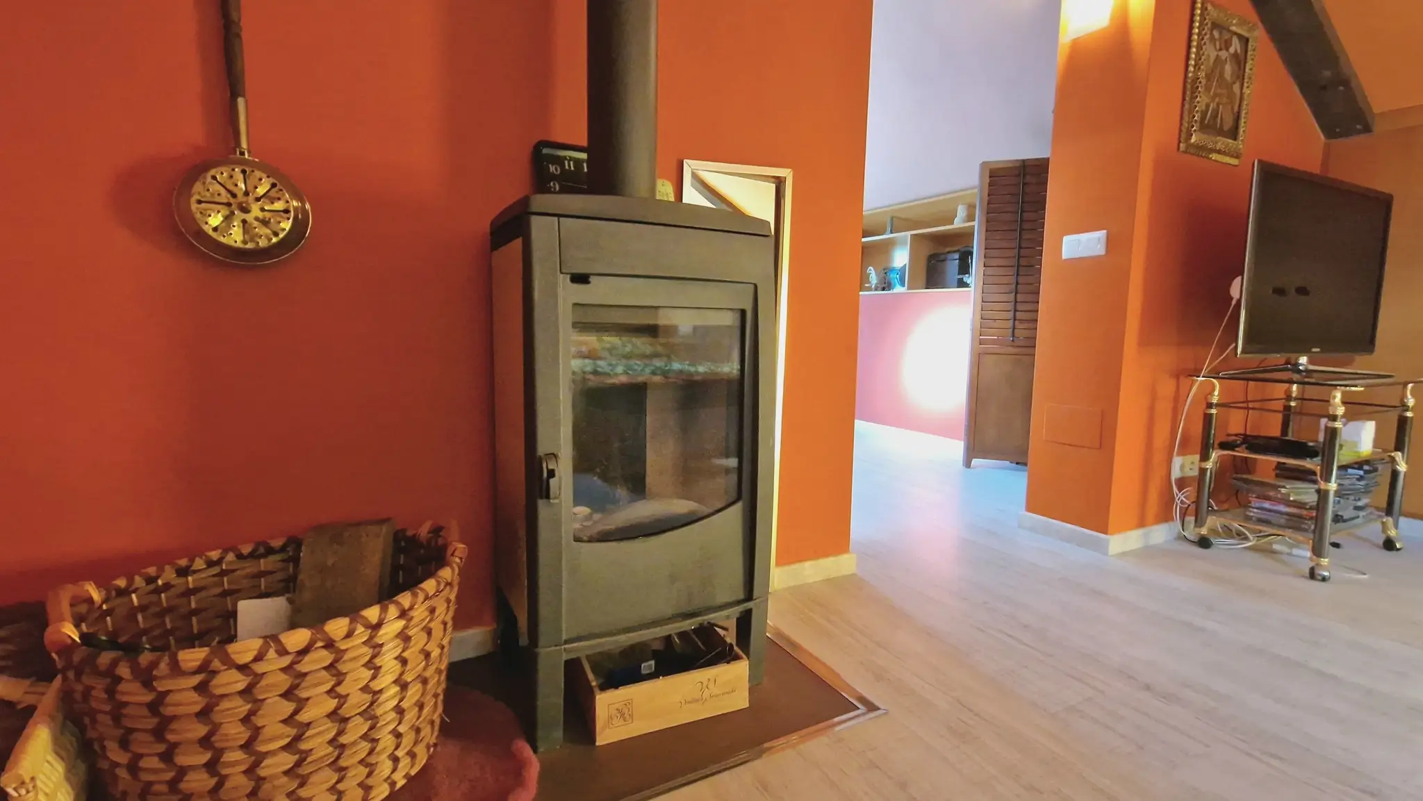 Poêle à bois moderne noir installé dans un salon aux murs terre cuite, avec un sol en bois clair et un panier à bûches traditionnel en osier.