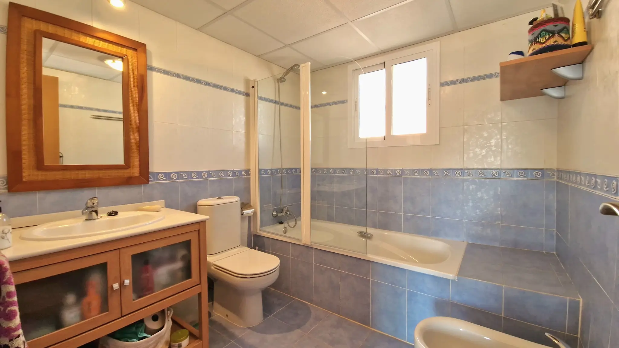 Salle de bain avec carrelage bleu et blanc, meuble sous-vasque en bois et baignoire avec paroi en verre, bénéficiant d'un éclairage naturel par une fenêtre.