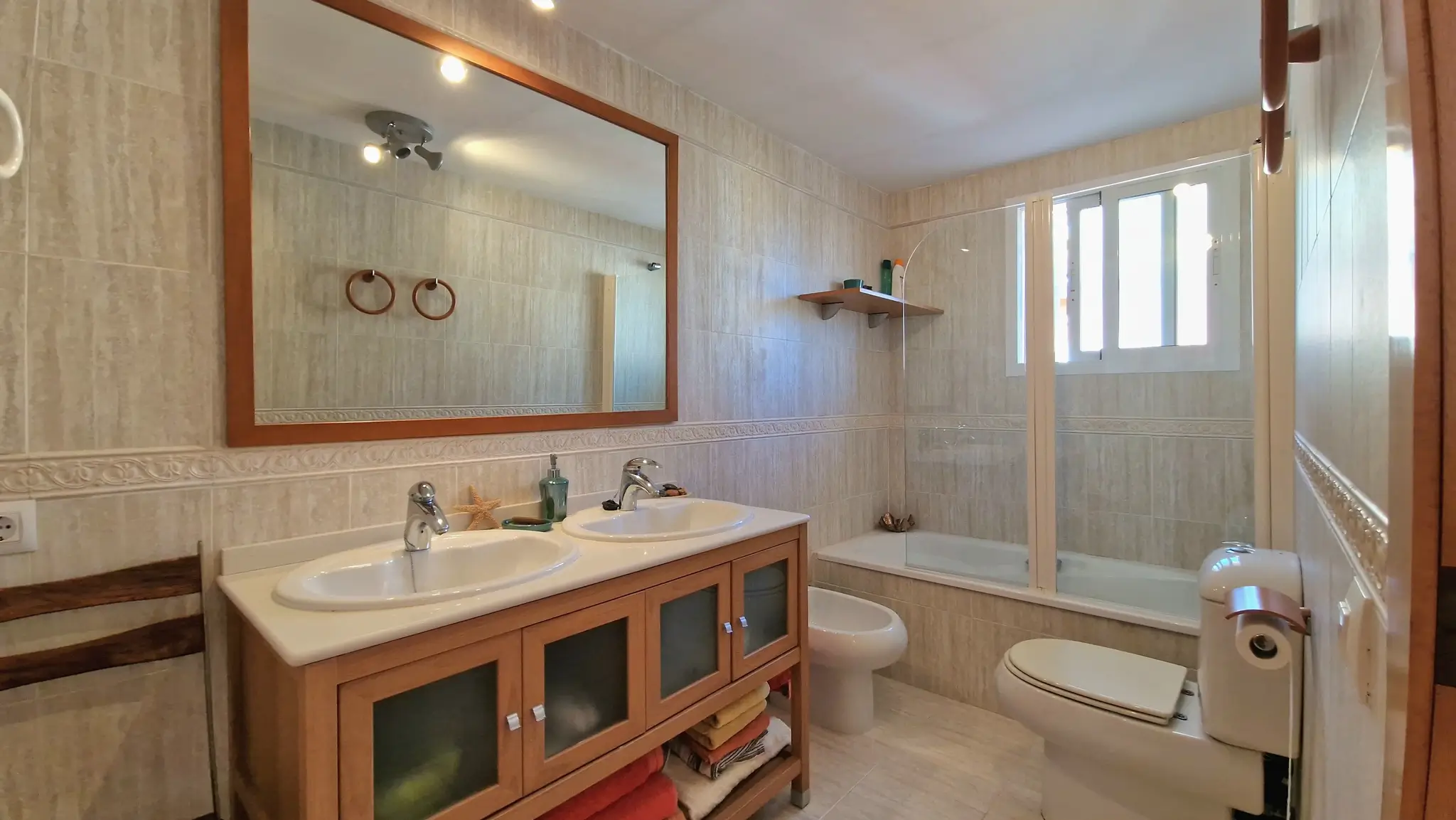 Salle de bain avec double vasque sur meuble en bois, grand miroir encadré, bidet et WC. Équipée d'une baignoire avec paroi vitrée et de murs carrelés aux tons naturels.