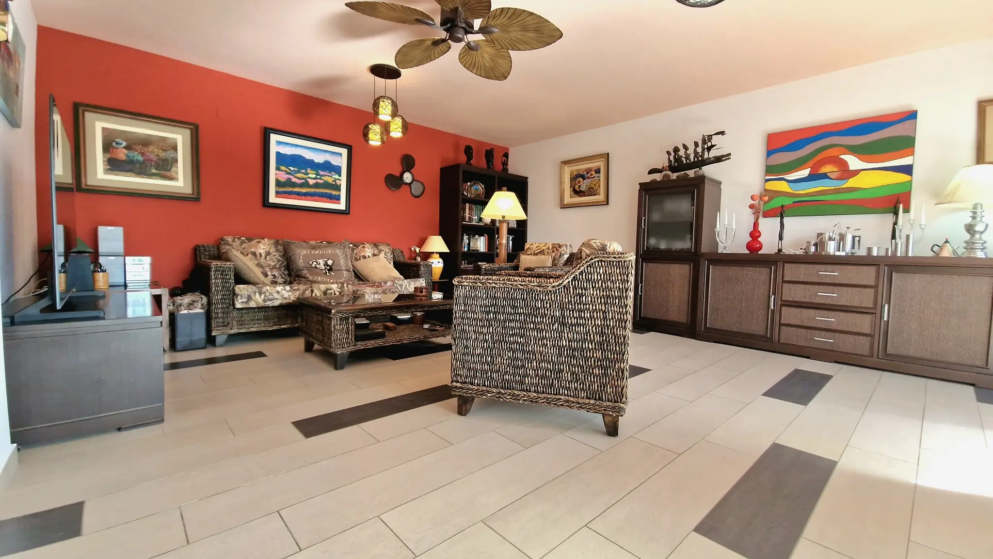 Salon avec mobilier en osier, mur d'accent terre cuite et ventilateur de plafond tropical. Le sol est revêtu de grands carreaux avec inserts décoratifs.