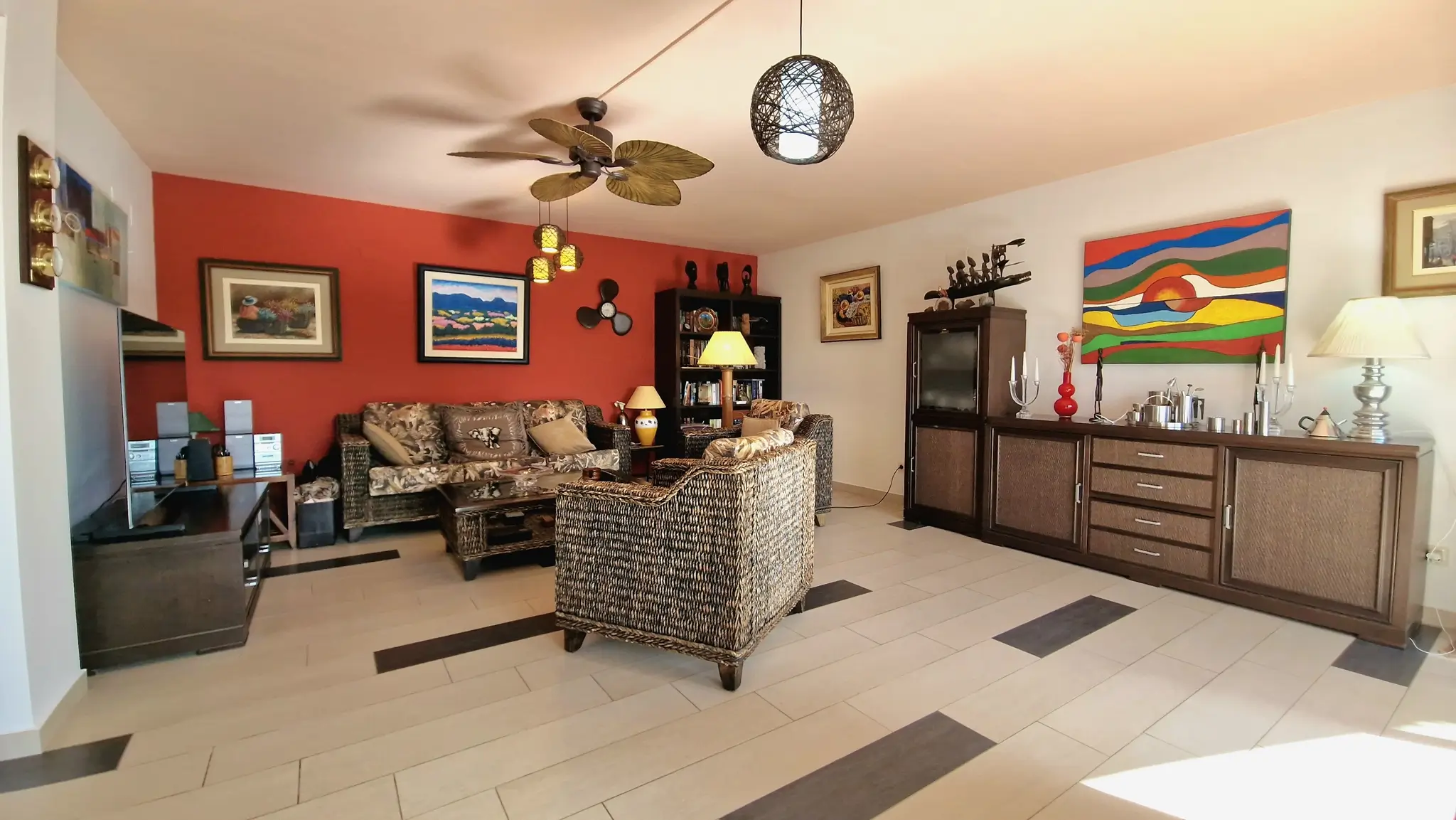 Salon avec un mur d'accent rouge, des meubles en osier et un ventilateur de plafond. Le sol est en carrelage clair avec des inserts sombres et des œuvres d'art variées.