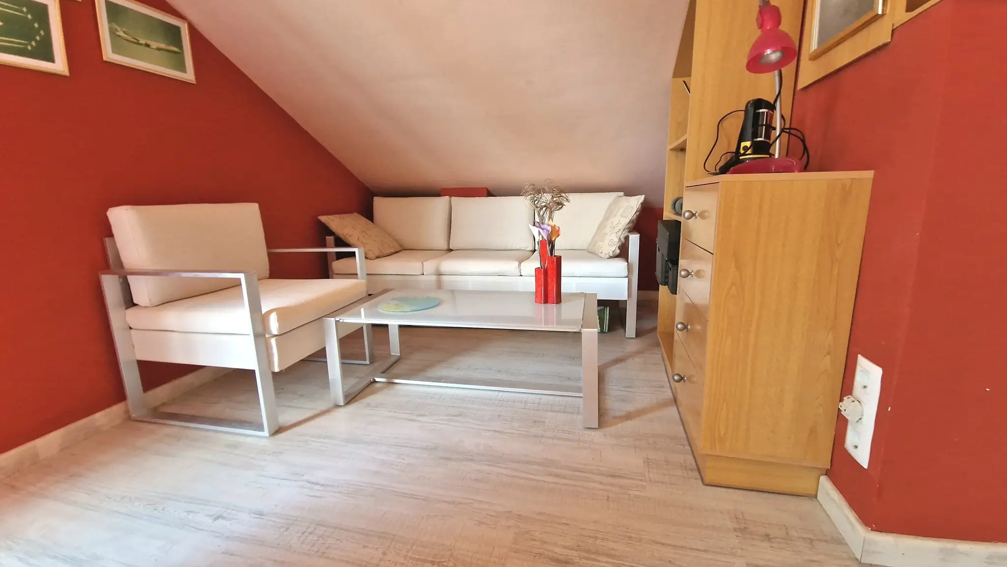 Salon mansardé avec murs rouges, canapé et fauteuil blancs à structure métallique, table basse en verre et sol clair.