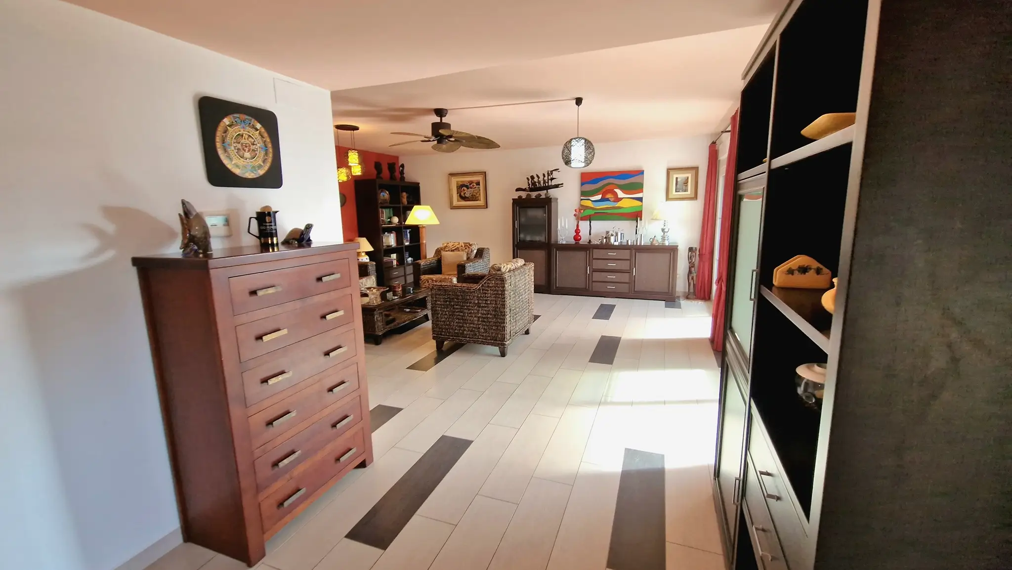 Salon spacieux avec carrelage clair à motifs sombres, fauteuils en osier, commode en bois massif et ventilateur de plafond de style colonial.