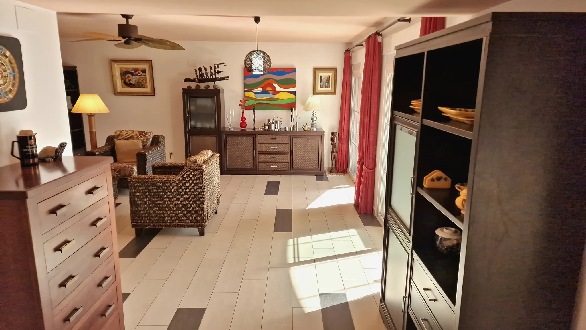 Salon spacieux avec fauteuils en osier, buffet en bois sombre et sol carrelé bicolore. Des rideaux rouges encadrent l'accès à la lumière naturelle.