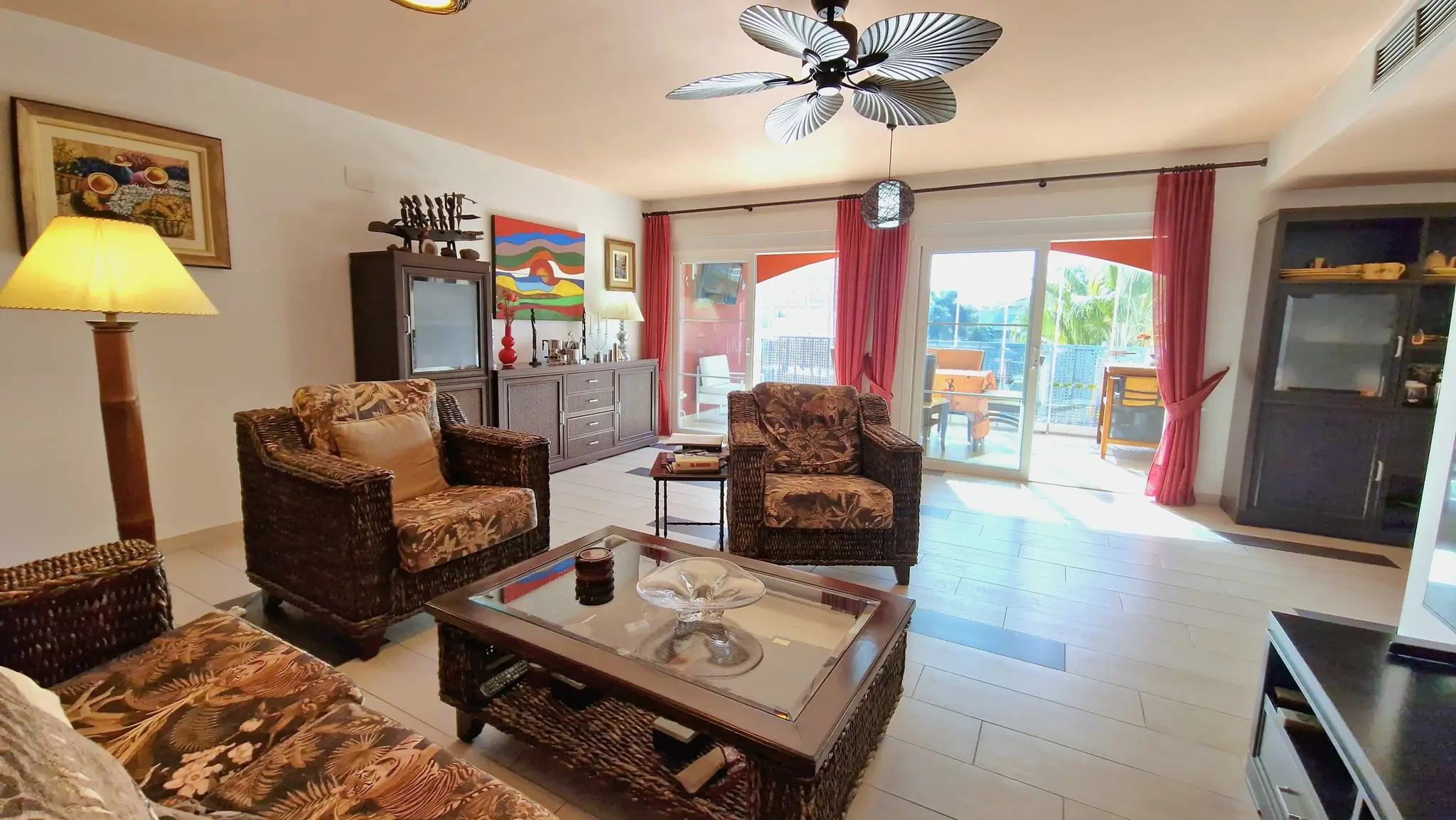 Salon spacieux avec mobilier en osier aux motifs tropicaux, ventilateur de plafond en forme de palmes et accès direct à la terrasse par des baies vitrées.