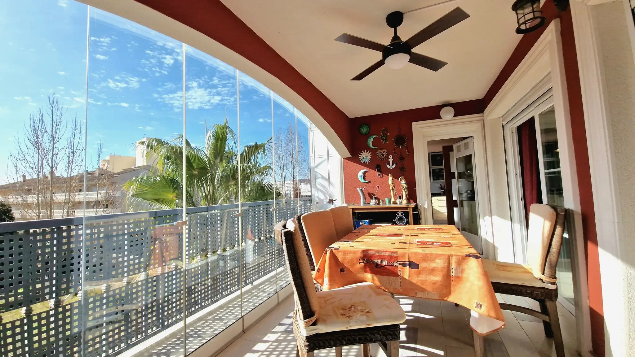 Terrasse couverte avec rideaux de verre sans cadre, coin repas extérieur avec chaises en osier, ventilateur de plafond et vue sur les palmiers.