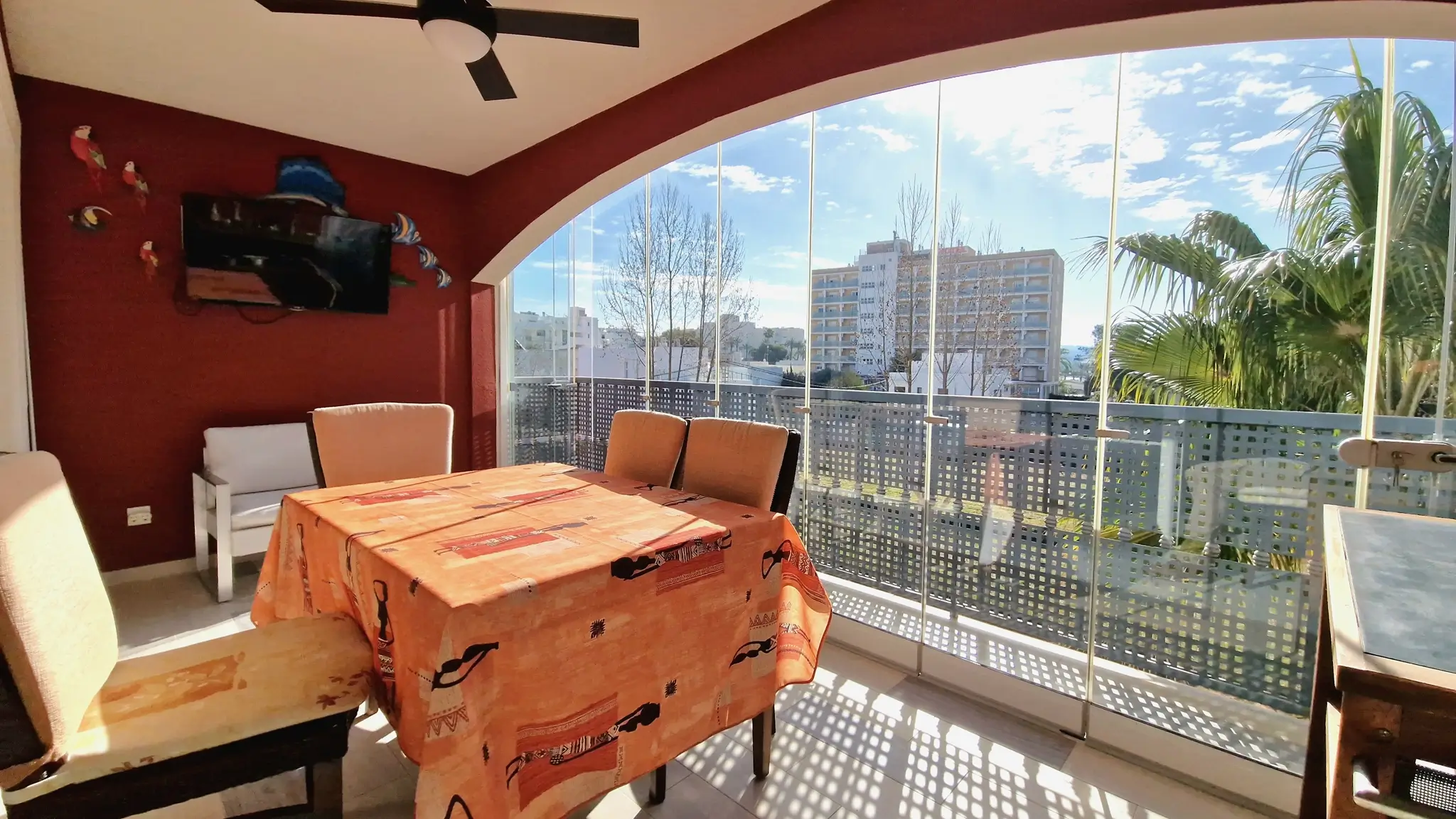 Terrasse fermée par des rideaux de verre, comprenant un coin repas, un mur rouge avec télévision et une vue dégagée sur la ville.