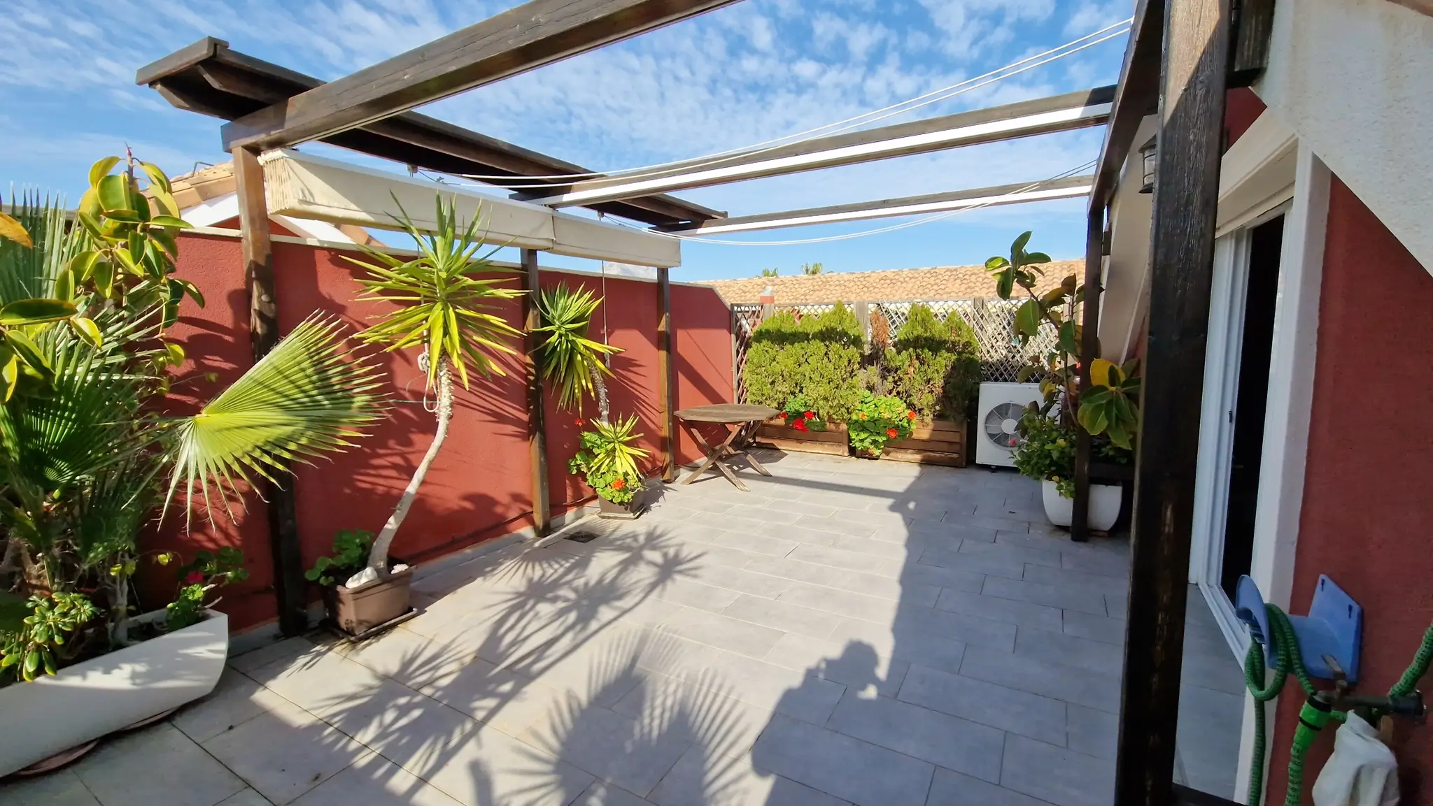 Terrasse privée avec carrelage gris, pergola en bois sombre et stores rétractables. Murs terracotta, palmiers en pots et petite table circulaire en bois sous le ciel bleu.