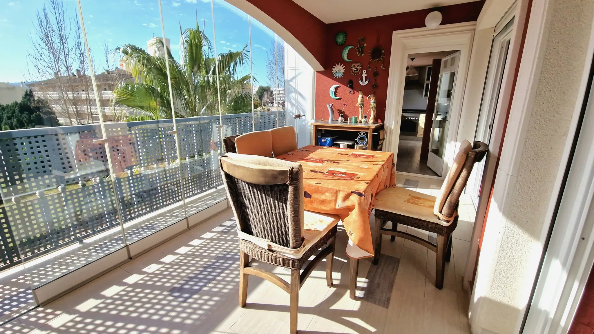 Terrasse vitrée avec rideaux de verre, espace repas extérieur et chaises en osier. Mur rouge décoré offrant une vue dégagée sur les palmiers.
