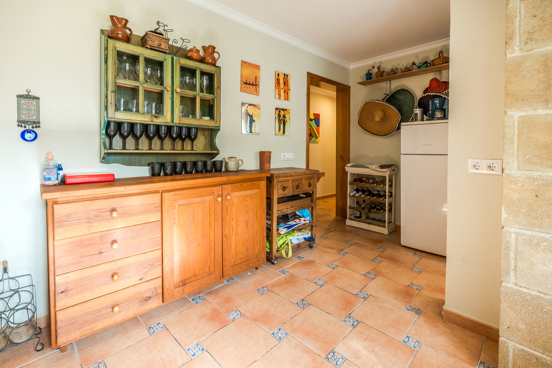 Buffet en bois rustique et vaisselier vert suspendu sur un sol en terre cuite avec inserts décoratifs. Un réfrigérateur blanc et un casier à vin complètent cet espace fonctionnel.