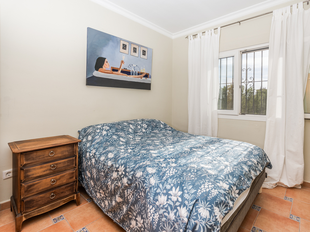 Chambre avec lit double, couette à motifs floraux bleus, table de chevet en bois et sol en terre cuite avec petits carreaux décoratifs.