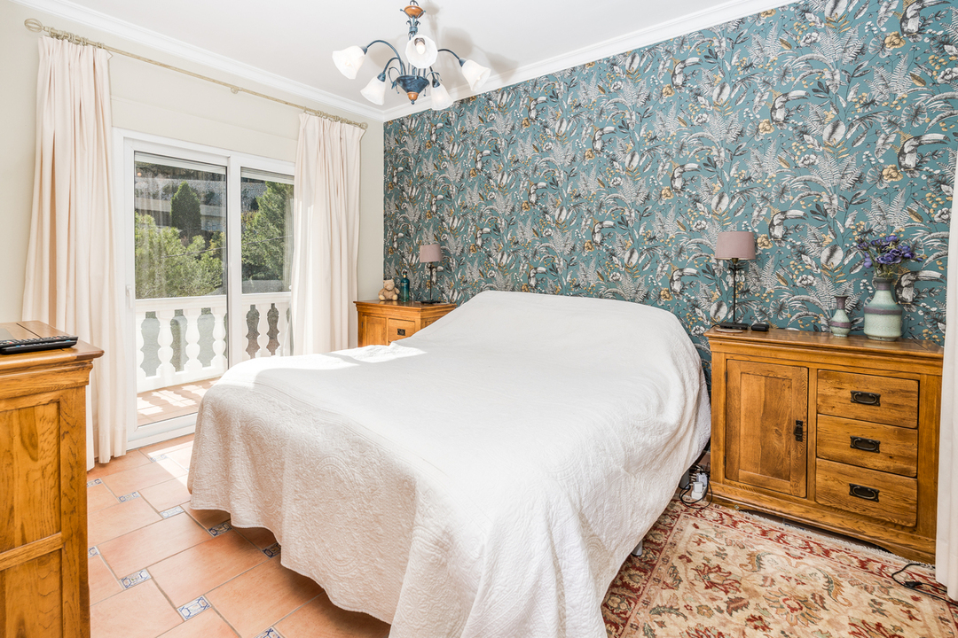 Chambre avec papier peint à motif floral, mobilier en bois et sol en terre cuite. Une porte vitrée coulissante mène à un balcon avec balustrade blanche.