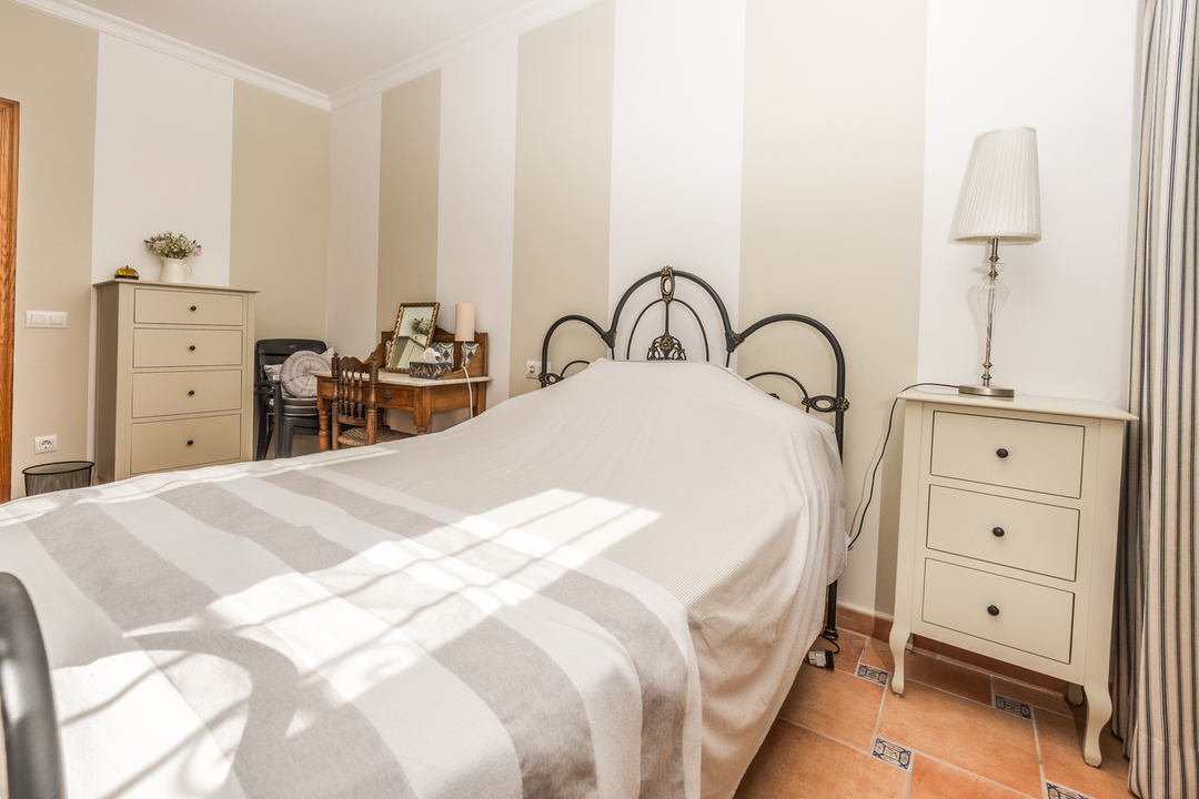 Chambre avec tête de lit en fer forgé, papier peint rayé et sol en terre cuite. Dispose d'un chevet crème et d'une coiffeuse en bois classique.