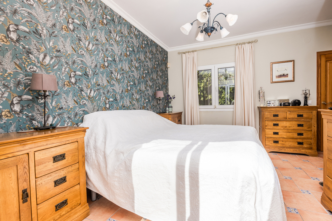 Chambre avec un mur d'accent au motif tropical, des commodes en bois massif et un sol en terre cuite orné de cabochons décoratifs. Éclairage par lustre classique.