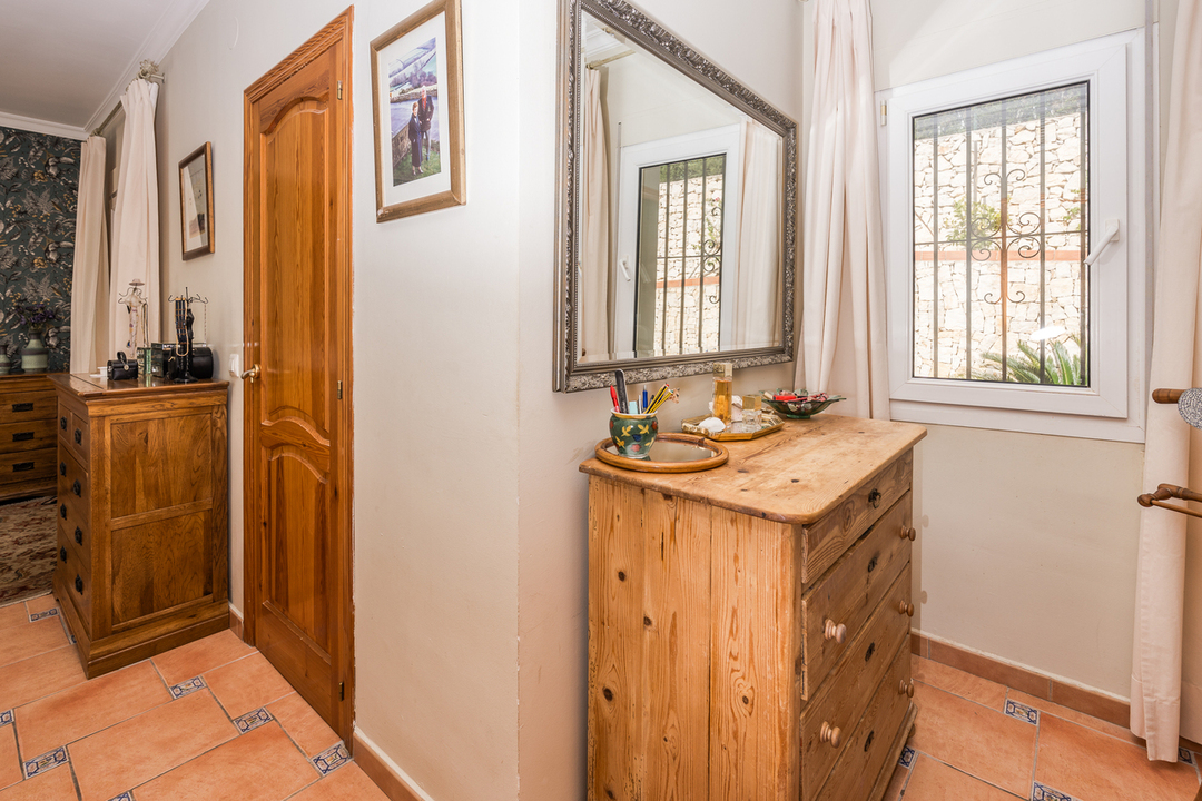Coin chambre avec commode en bois massif, sol en terre cuite aux inserts décoratifs et grand miroir à cadre argenté placé à côté d'une fenêtre.