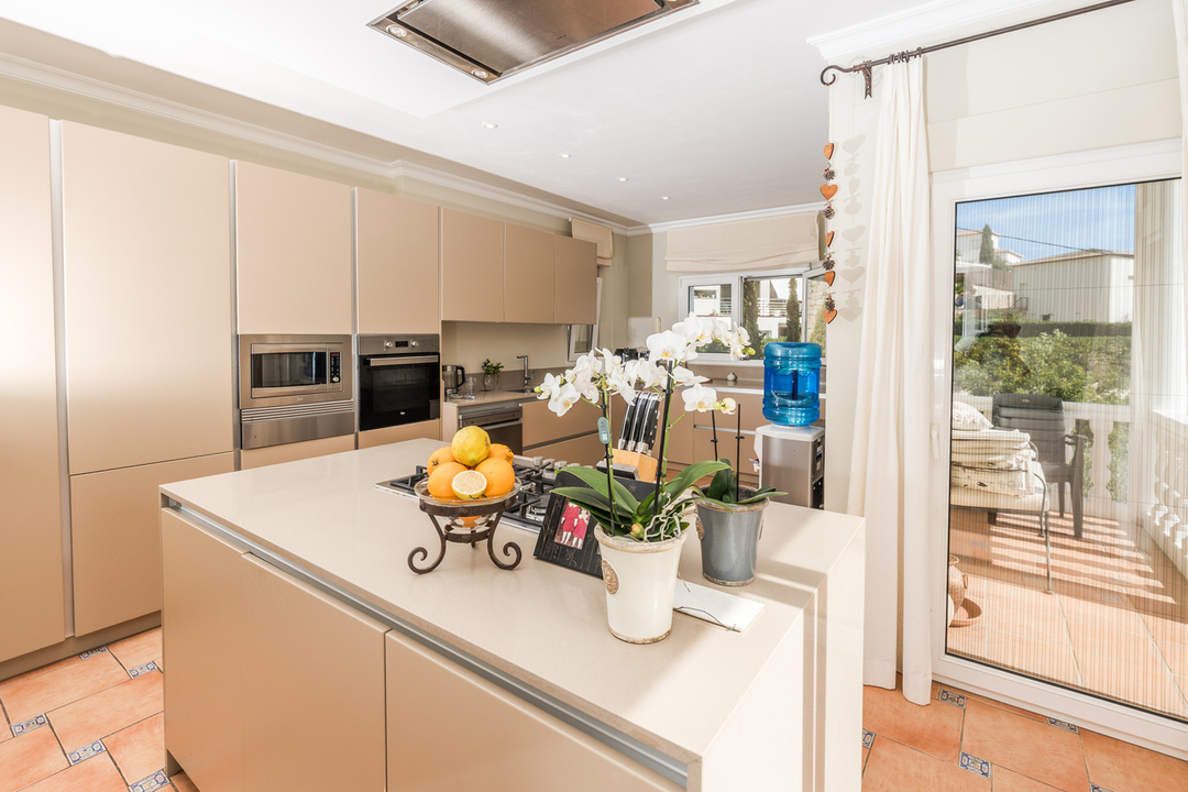 Cuisine moderne avec îlot central beige, plaque de cuisson à gaz, appareils intégrés et sol en terre cuite. Une porte vitrée offre un accès direct à la terrasse.