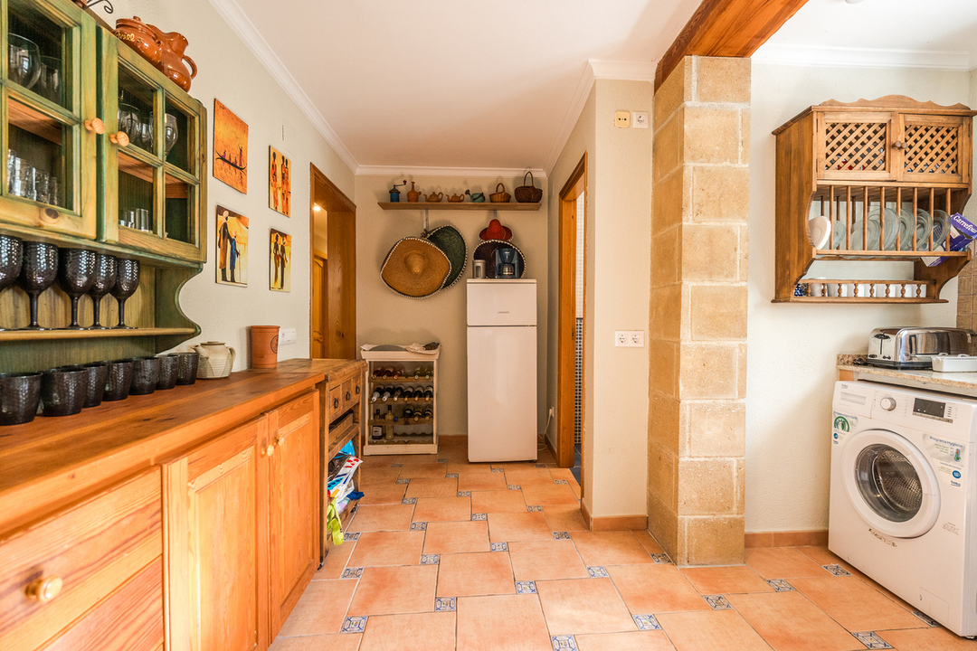 Cuisine rustique avec meubles en bois, sol en terre cuite à cabochons bleus, pilier en pierre naturelle et espace buanderie avec lave-linge.