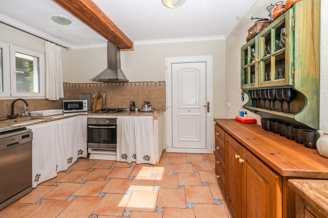 Cuisine rustique avec sol en terre cuite, poutres apparentes en bois, plans de travail en granit et buffet en bois avec vaisselier mural vert.