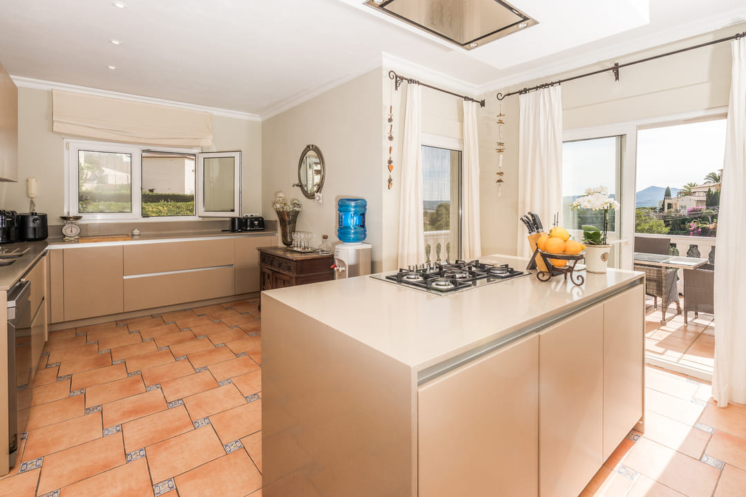 Cuisine spacieuse avec îlot central, plaques de cuisson au gaz, meubles beiges et sol en terre cuite. Les grandes ouvertures offrent une luminosité naturelle et un accès terrasse.