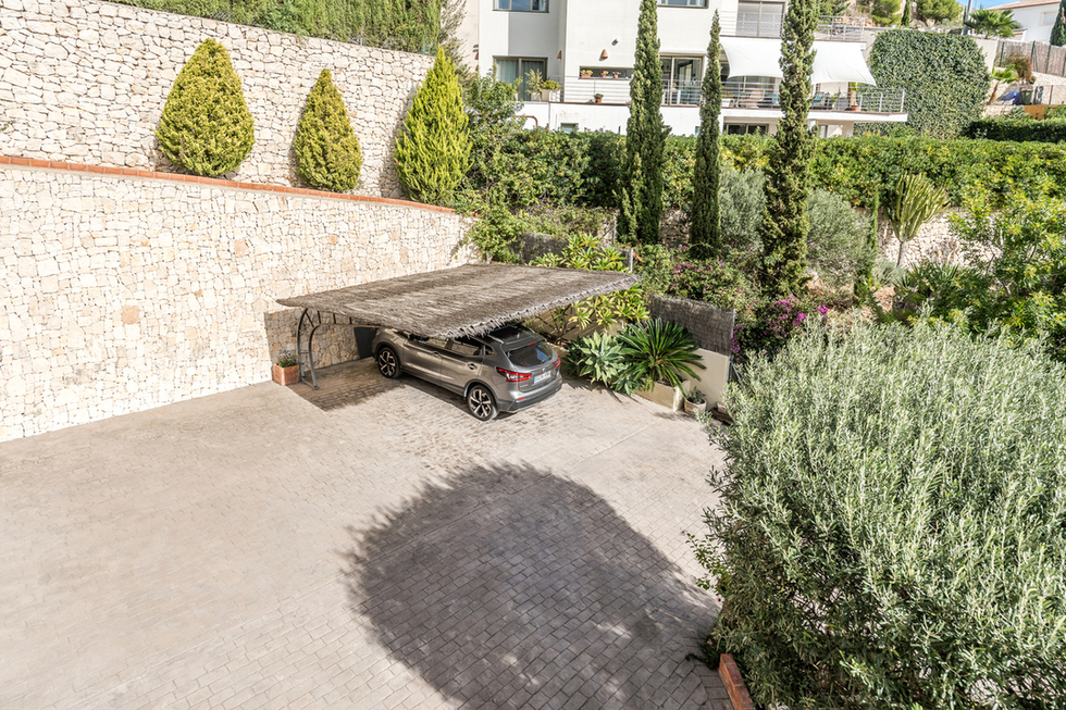 Espace de stationnement extérieur avec pavage au sol et abri en bruyère, bordé par un mur en pierre sèche et une végétation méditerranéenne.