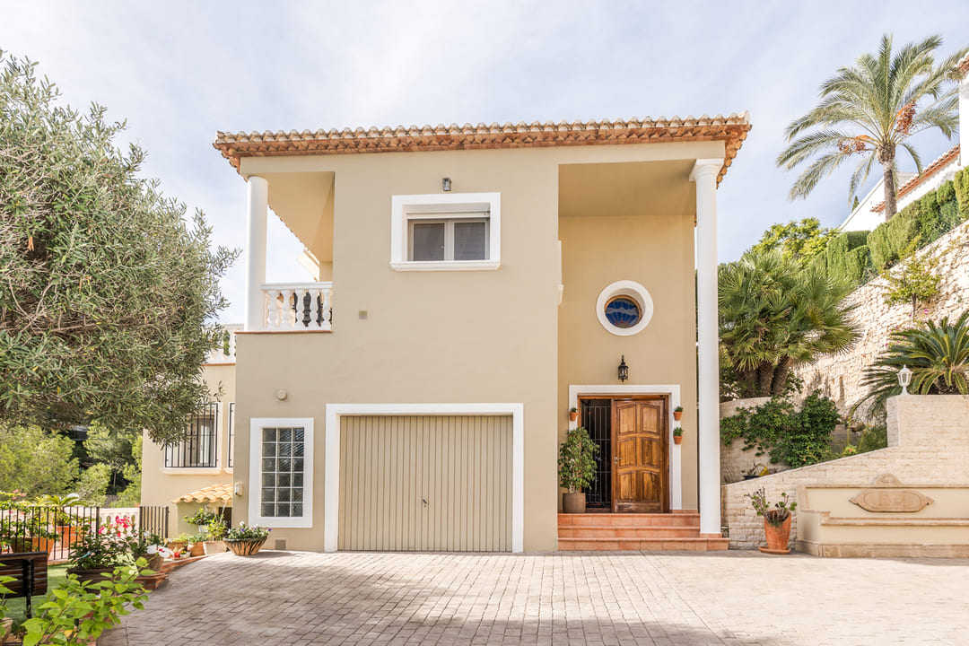 Façade extérieure d'une villa à deux étages avec finition beige, garage intégré, porte d'entrée en bois et allée pavée entourée de végétation méditerranéenne.