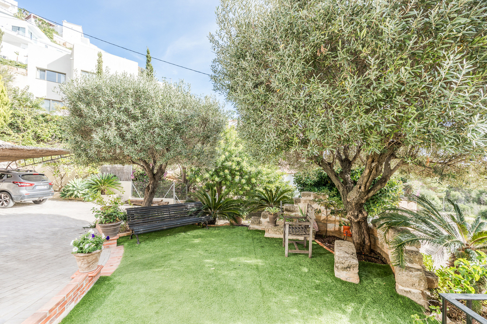 Jardin avec gazon synthétique, oliviers matures et banc en métal noir. Présence de murets en pierre et d'une zone de stationnement pavée et abritée.