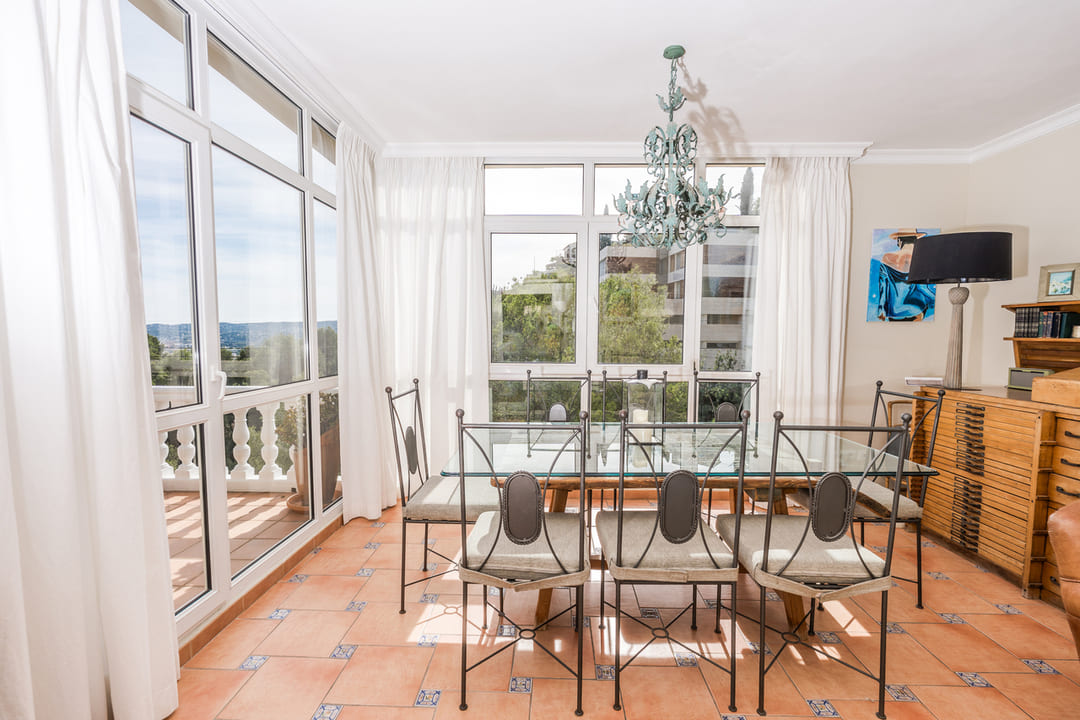 Salle à manger avec table en verre et chaises en fer forgé sur sol en terre cuite. De larges baies vitrées offrent une vue dégagée et un accès direct au balcon.