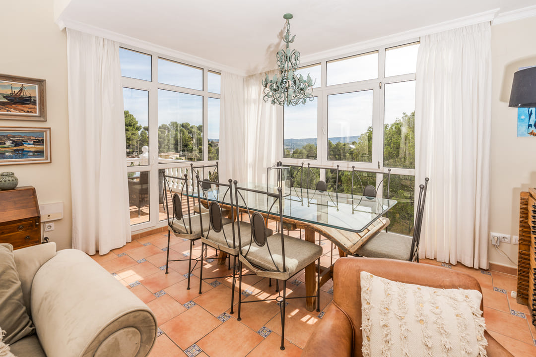Salle à manger lumineuse avec table en verre, chaises en fer forgé et sol en terre cuite. Grandes baies vitrées offrant une vue sur la végétation environnante.