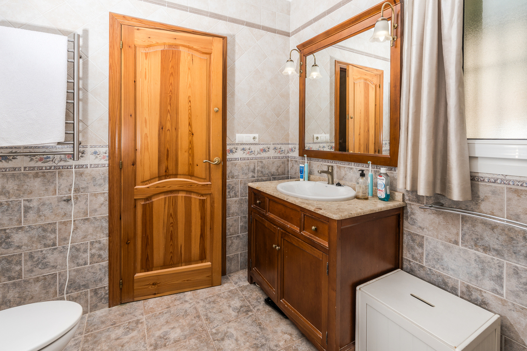 Salle de bain avec meuble vasque en bois, plan de travail en pierre et miroir assorti. Murs carrelés avec frise florale, porte en bois naturel et sèche-serviettes.