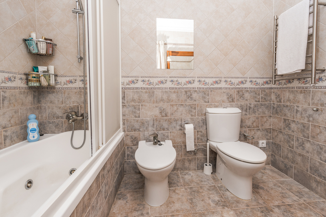 Salle de bain complète avec baignoire, bidet et toilettes. Les murs sont revêtus de carrelage aux tons terreux avec une frise florale et un sèche-serviettes chromé.
