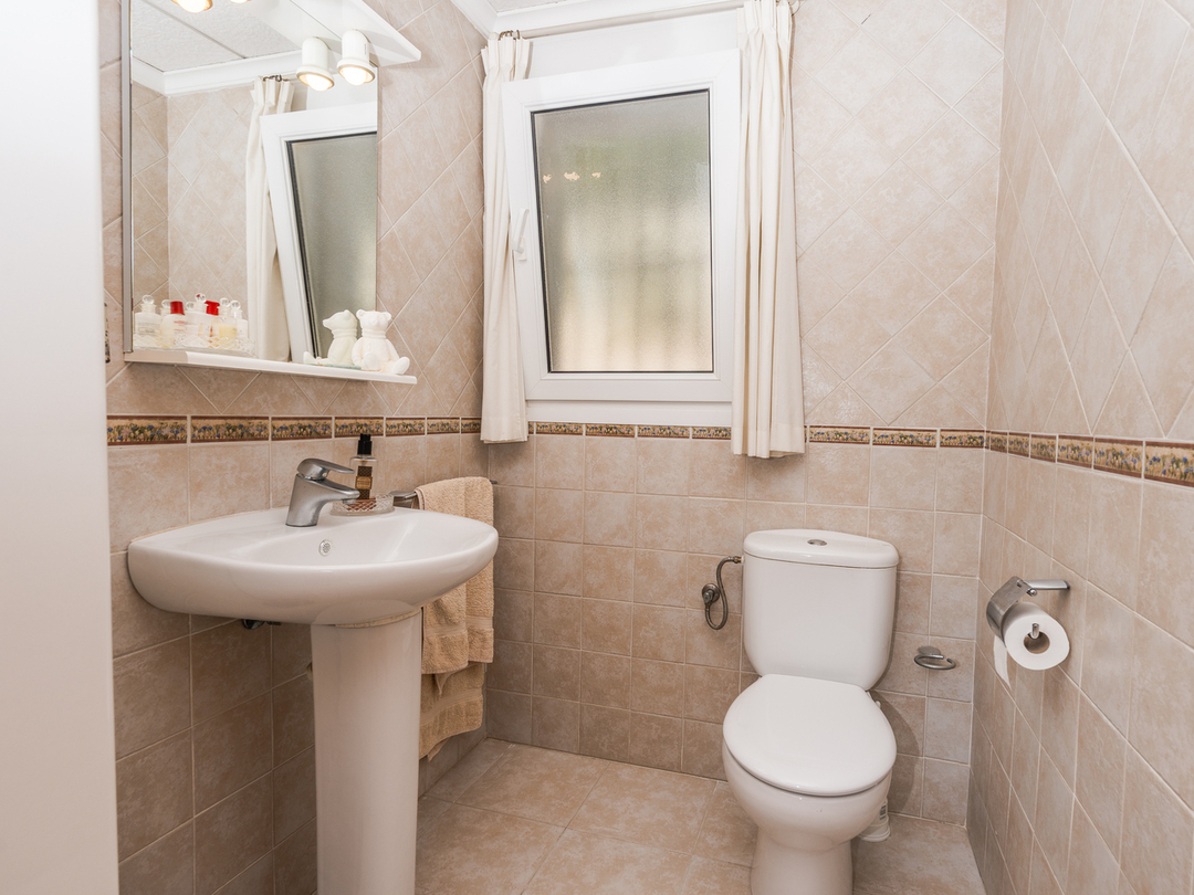 Salle de bains avec lavabo sur colonne, toilettes en céramique blanche et murs carrelés beige avec frise décorative et fenêtre dépolie.