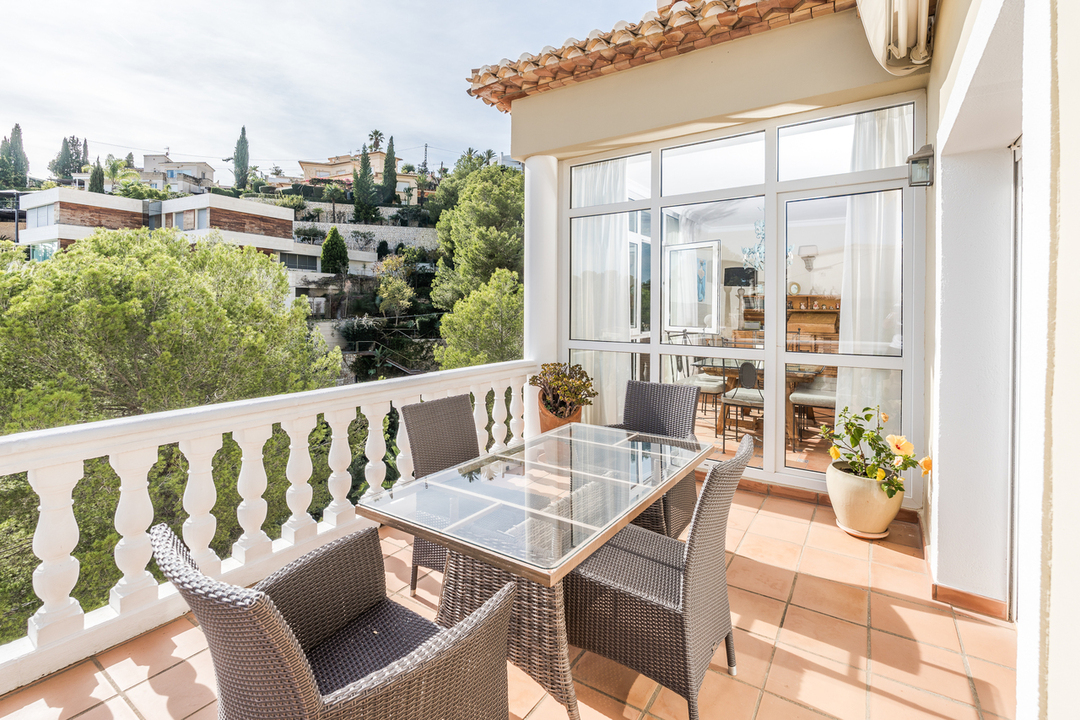 Terrasse avec carrelage en terre cuite, balustrade blanche et table en verre. Elle offre une vue sur la colline verdoyante et les résidences voisines.