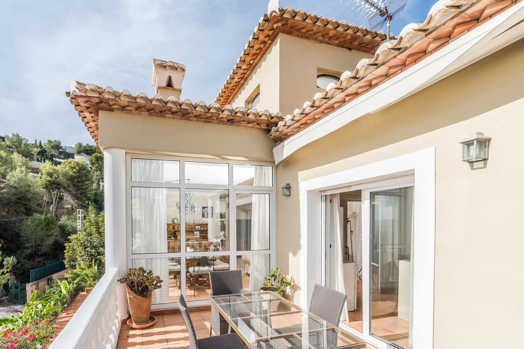 Terrasse en terre cuite avec table en verre et chaises en rotin. Balustrade blanche, verrière attenante et vue sur l'architecture méditerranéenne et les collines boisées.