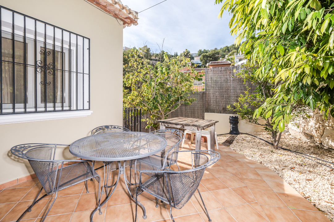 Terrasse extérieure avec carrelage en terre cuite, mobilier de jardin en fer forgé et fenêtre sécurisée, entourée d'arbres méditerranéens et d'un sol en gravier.