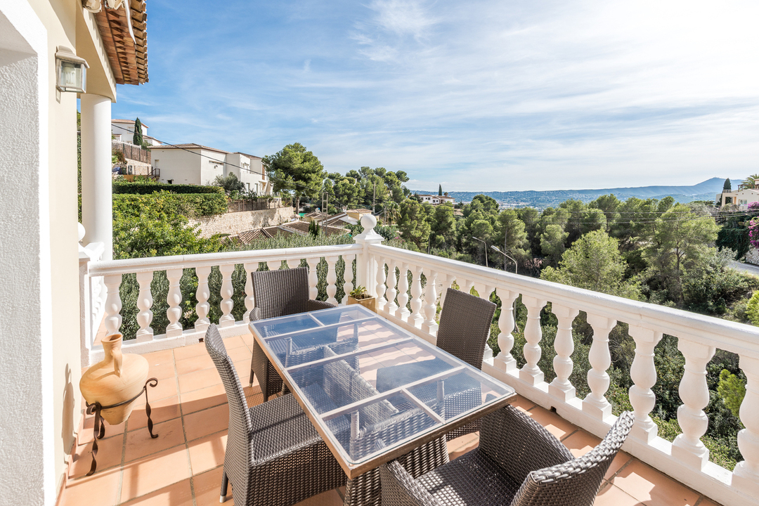 Terrasse surélevée avec table en verre, chaises en rotin et balustrade blanche, offrant une vue panoramique sur la vallée boisée et les collines.