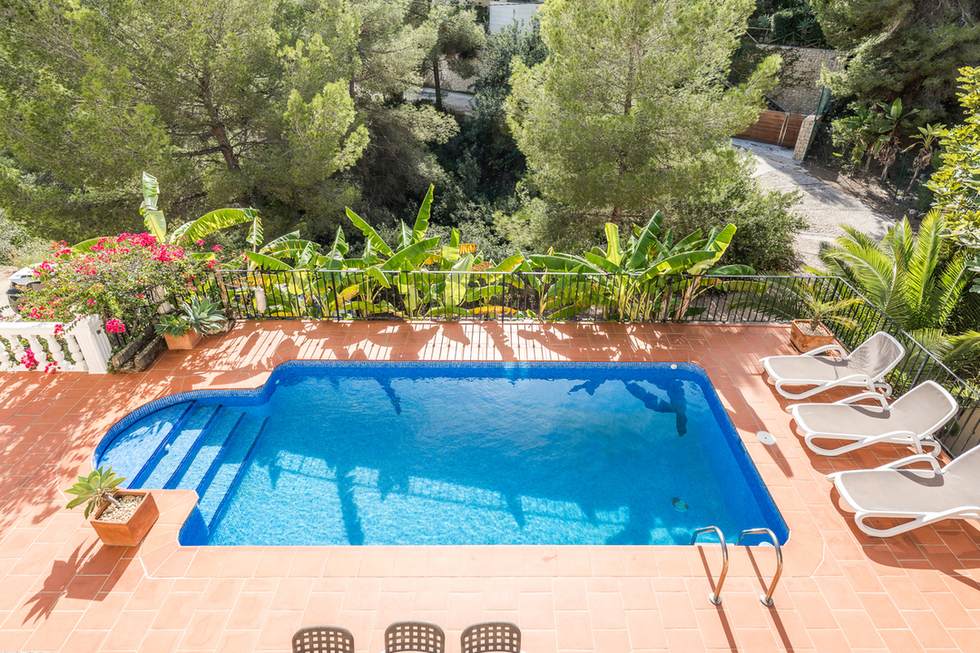 Vue en surplomb d'une piscine rectangulaire en mosaïque bleue avec terrasse en terre cuite, chaises longues et végétation méditerranéenne dense comprenant des pins.