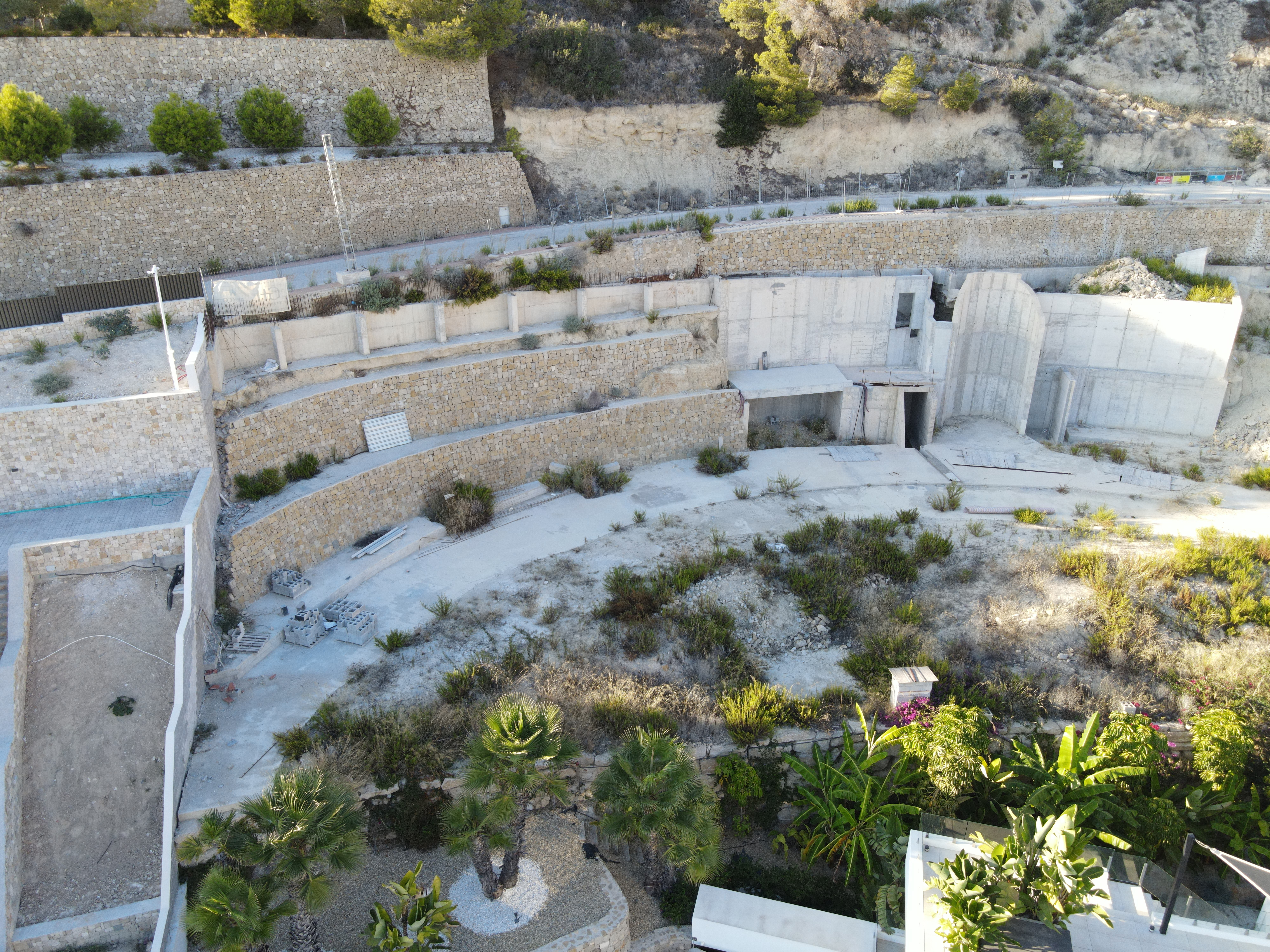 Vue aérienne de murs de soutènement en pierre et de structures en béton intégrés dans un terrain en pente avec des palmiers et arbustes.
